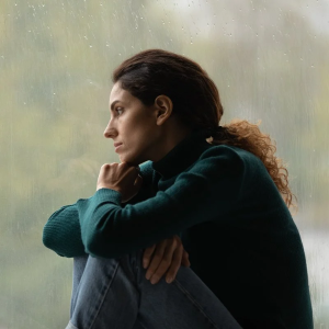 woman sits in front of window and gazes through the window to the rain outside