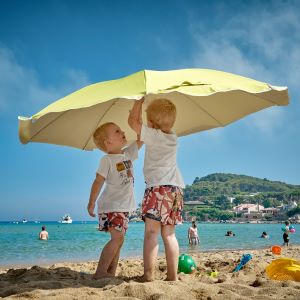 two young children surrounded by sand toys are putting up a beach umbrella whilst swimmers are bathing in the ocean under a bright blue sky