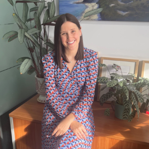 Smiling lady in a patterned dress sitting on the edge of a table with plants in the background