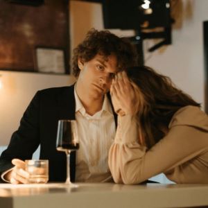 A couple are sitting at a bench.  The woman has a glass of wine and holds her head in her hands, whilst the man holding a whisky looks past the woman with a bored expression