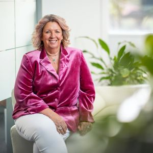 Suzanne Jones smiling at the camera, sitting on the arm of a couch wearing a pink jacket and white pants 